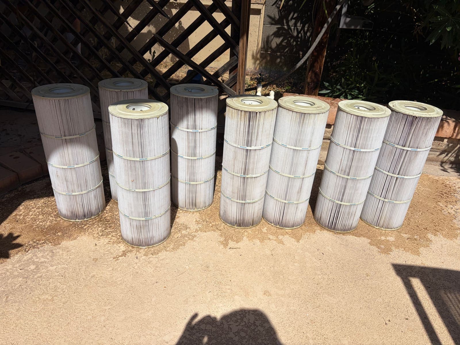 Eight pool filter cartridges lined up on a deck mid-cleaning — the washed ones white, the rest still gray with Arizona dust